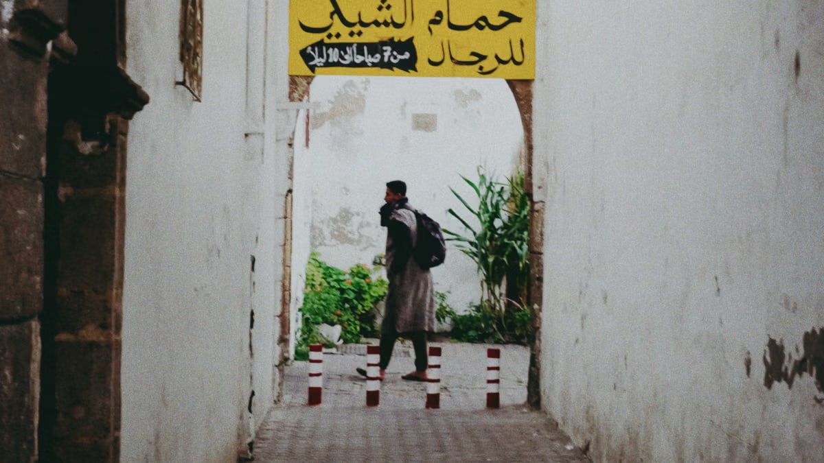 Man walking through a rustic alleyway with Arabic signage, surrounded by white walls and vegetation.
